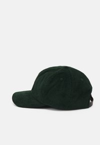 Grønn kordfløyelslue med buet skygge, synlig ribbestruktur, justerbar stropp bak og ventilasjonshull.