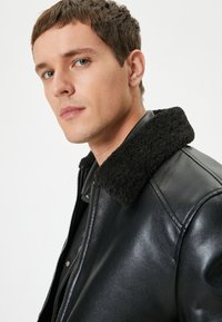 Chaqueta de cuero negra con un cuello de felpa negra, cierre delantero con cremallera y textura suave. Presenta un diseño ajustado y mínimas adornos.