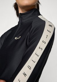 Schwarzes sportliches Quarter-Zip-Oberteil aus strukturiertem Material. Mit einem hellen Streifen auf der Schulter, auf dem "ENDLESS" in Schwarz gedruckt ist.
