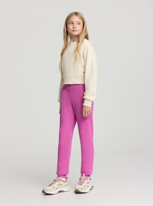 Felpa color crema in stile uncinetto, aderente in vita, abbinata a pantaloni della tuta rosa acceso con vita elasticizzata. Sneakers beige e rosa.