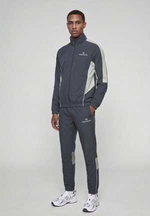 Homme debout portant un survêtement Sergio Tacchini gris foncé avec des panneaux gris clair et des baskets blanches New Balance, mains dans les poches, fond neutre.