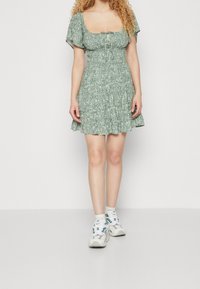 Osoba s kudrnatými vlasy nosí krátké zelené květinové šaty a bílé tenisky, stojí proti jednobarevnému bílému pozadí.