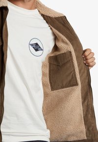 Chaqueta marrón con cremallera y forro de felpa, que revela una camiseta blanca con un logotipo gráfico. Incluye un bolsillo de pana marrón en el interior.