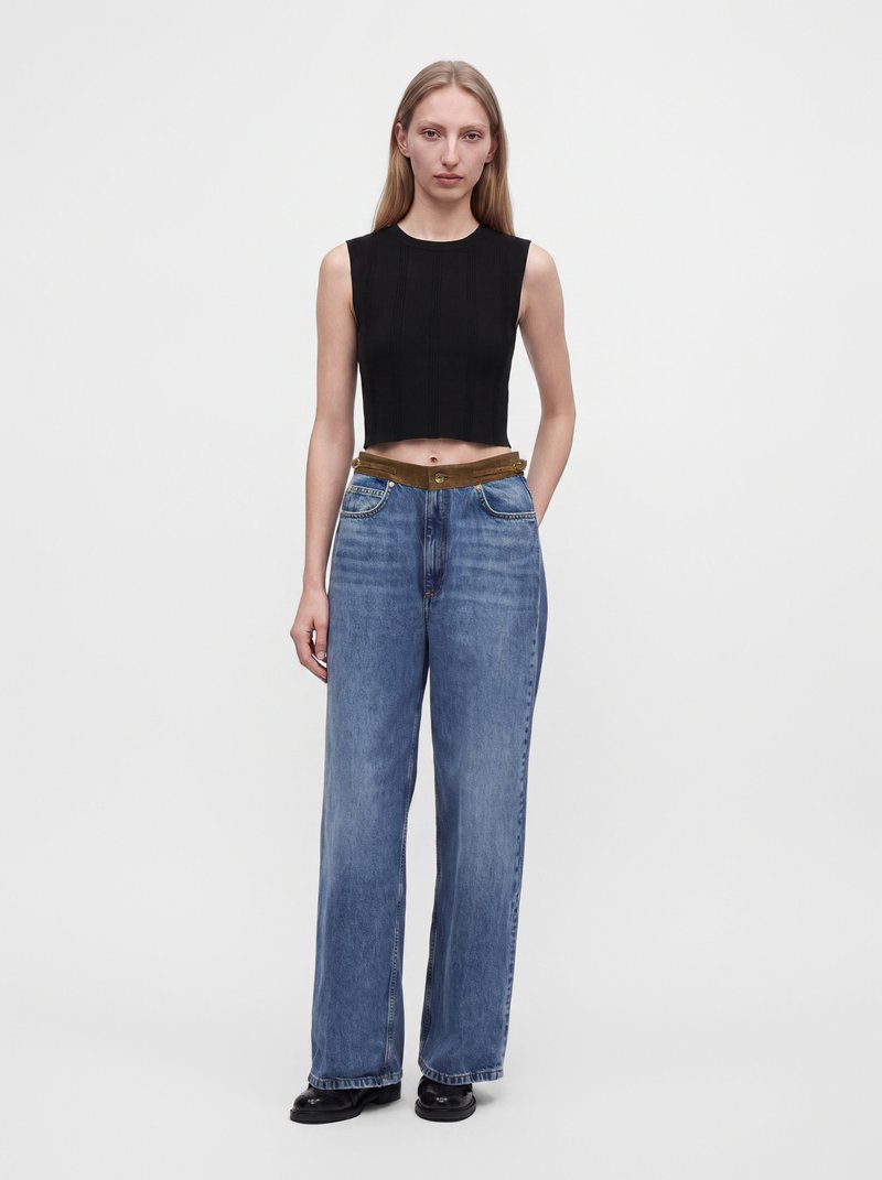 Jeune femme aux longs cheveux blonds portant un crop top noir sans manches, un jean bleu taille haute à jambes larges, et des chaussures noires, debout sur un fond blanc.