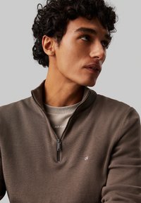 Brauner Reißverschluss-Sweatshirt aus strukturiertem Stoff, mit einem gestickten Logo auf der Brust, getragen über einem beigen Hemd.