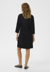 Vestido de mezclilla negro con cintura ajustada, mangas tres cuartos y dobladillo recto, visto desde la parte de atrás, combinado con zapatos negros con detalles en plata.