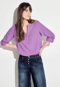 Blusa texturizada morada con escote abotonado, combinada con jeans de mezclilla oscuros de tiro alto que presentan múltiples botones frontales y bolsillos.