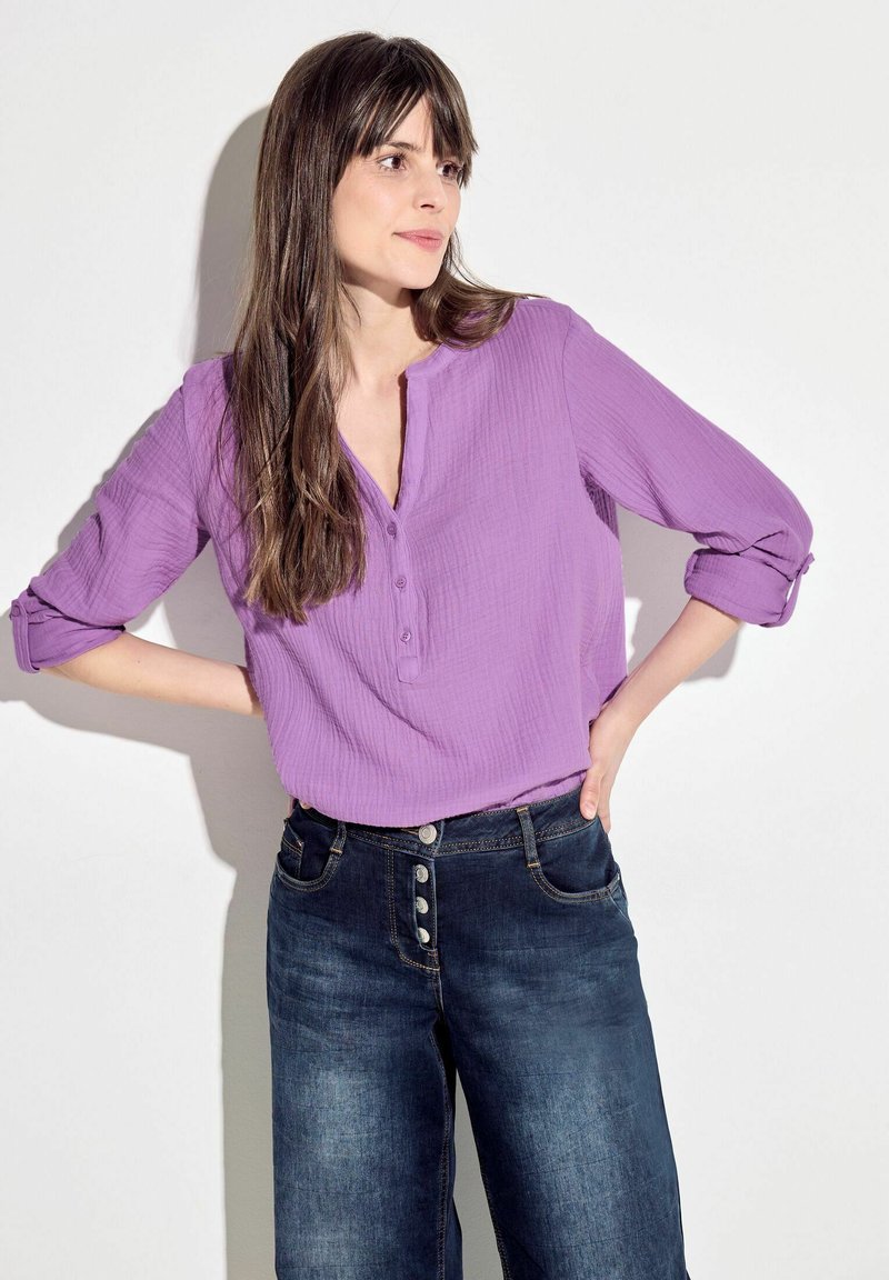 Blusa viola con texture e scollatura a bottoni, abbinata a jeans scuri a vita alta con vari bottoni e tasche frontali.