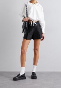 Sorte shorts med en glat tekstur, snoreliv og nitterdetaljer, parret med en hvid skjorte og sorte loafers med tykke såler.