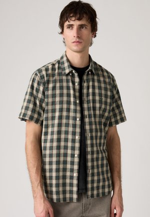 Uomo con camicia a maniche corte a quadri verde e beige sopra una t-shirt nera su sfondo uniforme.