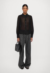Zwarte sheer blouse met textuur in de vorm van stippen, knopenfront en lange mouwen. Gepaarde met grijze wijde broek en een kleine zwarte handtas.