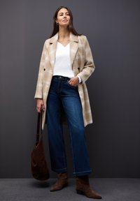 Beige karierter Wollmantel, weißes V-Ausschnitt-Oberteil, dunkelblaue weite Jeans, braune Wildleder-Stiefeletten und eine braune Umhängetasche. Einfaches Design, warme Farbtöne.