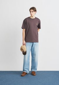 Νέος άνδρας που φοράει σκούρο μωβ μπλουζάκι και ανοιχτόχρωμα φαρδιά τζιν κρατάει καπελάκι μπεζ, στέκεται σε μπλε χαλί μπροστά σε λευκό τοίχο.