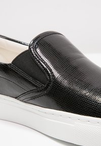 Chaussures noires à enfiler avec une surface texturée et brillante, des empiècements latéraux élastiques, une semelle en caoutchouc blanche et un design minimaliste.