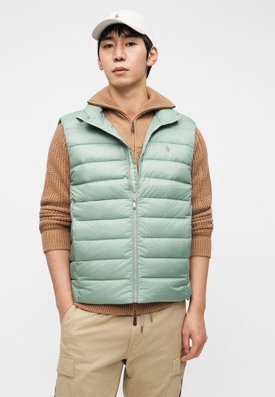 Ανοιχτό πράσινο αμάνικο μπουφάν με φερμουάρ μπροστά και ψηλό γιακά. Φορεμένο πάνω από ένα καφέ πλεκτό πουλόβερ, συνδυασμένο με μπεζ παντελόνι cargo.