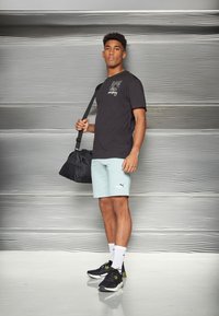 Camiseta gráfica preta, calções azul claro com o logo da Puma, mala de desporto preta, sapatos de atletismo pretos e verdes, em pé contra um fundo cinza texturizado.