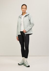 Ljusgrön vattentät jacka med dragkedja fram och huva, kombinerad med svarta leggings och ljusgrå sneakers. Har en texturerad design.