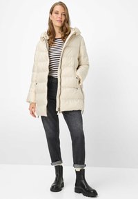 Flødefarvet dunjakke med hætte, der har et quiltet design og sidelommer. Bæres over en navy- og hvidstribet top, kombineret med mørke jeans og sorte støvler.