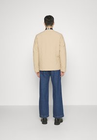 Beige gesteppte Jacke mit geradem Schnitt, kombiniert mit blauen weit geschnittenen Jeans und schwarzen Schuhen, von hinten betrachtet, zeigt ein Rautenmuster.
