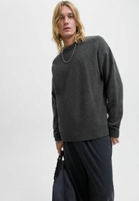 Mørk grå strikket sweater med ribbet rund hals og mansjetter. Materialet ser blødt og struktureret ud, bæres med mørke bukser og en taske.