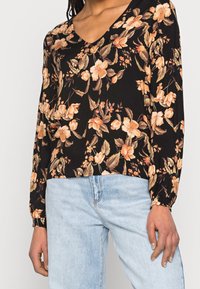 Blusa nera a maniche lunghe con motivo floreale in tonalità di pesca e crema, dotata di scollatura a V e polsini arricciati. Abbinata a un jeans denim blu chiaro.