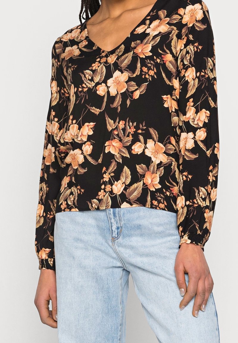 Blusa nera a maniche lunghe con motivo floreale in tonalità di pesca e crema, dotata di scollatura a V e polsini arricciati. Abbinata a un jeans denim blu chiaro.