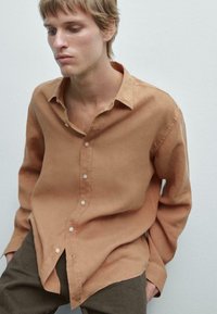Chemise marron à manches longues en tissu léger avec une coupe décontractée, dotée d'un col, d'une fermeture à boutons et d'un subtil éclat. Associée à un pantalon sombre.