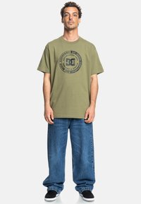 Olivegrünes T-Shirt mit kreisförmigem DC-Logo-Design, kombiniert mit weit geschnittenen blauen Jeans und schwarzen Skate-Schuhe.