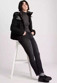 Czarna kurtka puchowa z błyszczącym kołnierzem i kapturem, szary golf, ciemne dżinsy i czarne obuwie, siedząca na białym stołku na neutralnym tle.