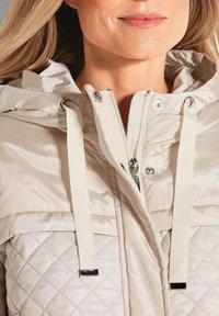 Hellbeige gepolsterte Jacke mit Kapuze, Reißverschluss und kontrastierenden glatten sowie gesteppten Texturen. Silberne Hardware-Akzente vervollständigen das Design.