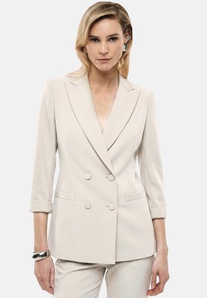 Vrouw met blond haar die een crème double-breasted blazer en bijpassende broek draagt, met een zilveren armband en opvallende oorbellen.