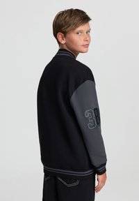 Czarna i szara kurtka varsity z prążkowanym kołnierzem i poziomymi paskami przy mankietach. Posiada przerośnięty numer 38 na szarym rękawie.