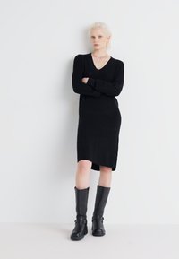 Black long-sleeve ribbed dress with a deep V-neck and knee-length hem, paired with tall black leather boots featuring buckle accents.