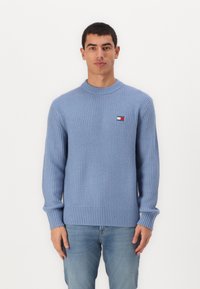 Lichtblauwe ribgebreide trui met een ronde hals en lange mouwen. Bevat een klein logopatch op de borst. G gedragen over blauwe spijkerbroek.