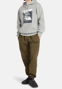 Grå huvtröja med grafiskt tryck och dragsko, i kombination med löst sittande olivgröna byxor och svarta slip-on skor.