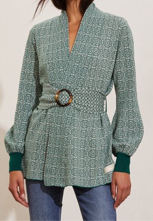 Grønblomstret bluse med indsvungen design, samlet om taljen med et skildpadde-mønsterbælte. Har puffede ærmer og ribbede manchetter.