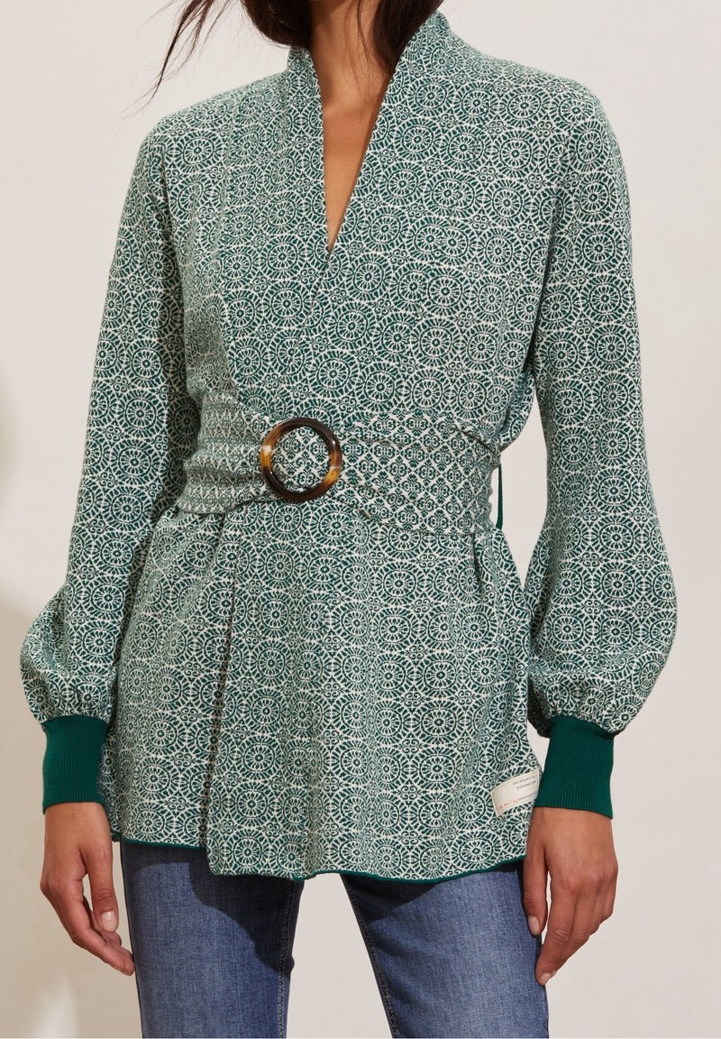 Vihreä kuvioitu pusero, jossa on kietaisumalli ja joka on vyötetty kääntökuvioisella vyöllä vyötäröltä. Sisältää puffimaiset hihat ja ribattu hihansuu.