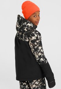 Schwarze und gemusterte Daunenjacke mit Kapuze; weist ein Spritzdesign in Creme und Braun im oberen Bereich auf, hat ein strukturiertes Gewebe und wird mit einer orangefarbenen Mütze kombiniert.