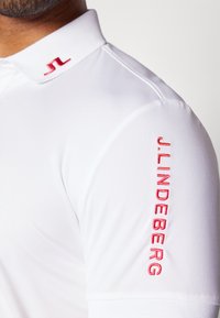 Vit polo skjorta med en strukturerad krage. Har en röd broderad logotyp på kragen och texten "J. Lindeberg" på ärmen. Slät textur.