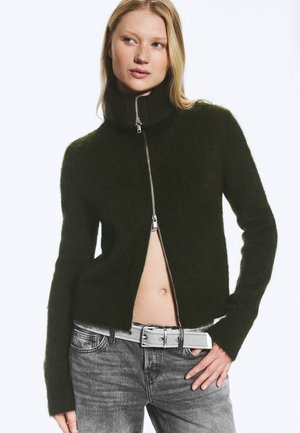 Femme portant un pull zippé duveteux vert foncé, partiellement ouvert, montrant le ventre, avec un jean gris et une ceinture blanche, posant avec la main sur la hanche.