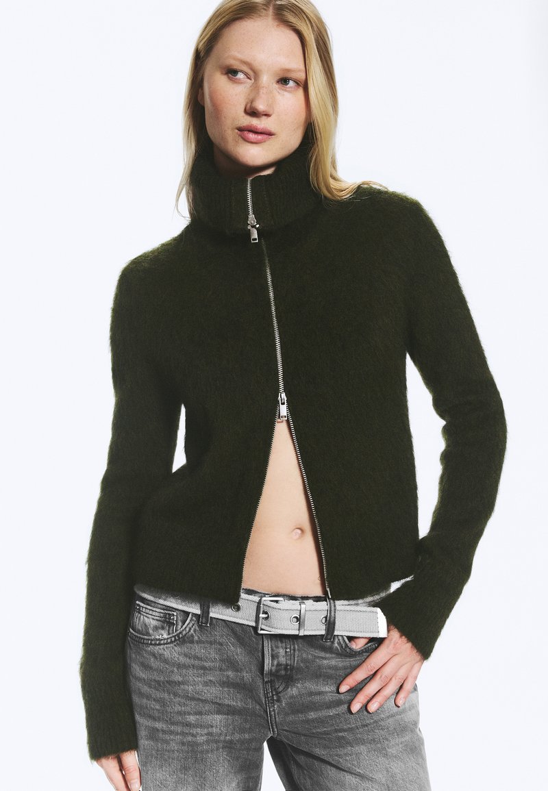 Femme portant un pull zippé duveteux vert foncé, partiellement ouvert, montrant le ventre, avec un jean gris et une ceinture blanche, posant avec la main sur la hanche.