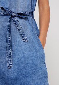 Jeansové šaty světle modré barvy s páskem v oblasti pasu a bočními kapsami. Tkanina má hladkou strukturu s jemnými zesvětlenými detaily.