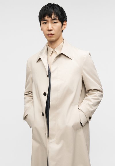 Μπεζ παλτό τύπου τρεντσότ με κλασικό γιακά, τρία σκουρόχρωμα κουμπιά και τσέπες στο πλάι, φορεμένο πάνω από ελαφρύ πουκάμισο και γκρι ύφασμα. Λεια υφή.