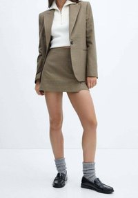 Blazer à carreaux marron avec mini jupe assortie, pull zippé blanc, chaussettes grises retroussées et mocassins noirs. Tissu doux et design ajusté.