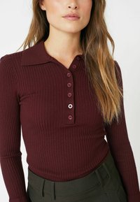 Burgundy ribbed polo shirt with a collar, featuring a button-up placket and long sleeves. Fabric is fitted and textured.