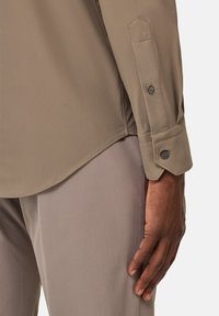 Camisa beige de manga larga con una textura suave, que presenta un puño abotonado y un dobladillo curvo, combinada con pantalones beige claros.