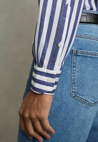 Camisa de rayas de manga larga en blanco y azul marino, con puños abotonados y un bolsillo, combinada con jeans de mezclilla azul claro.