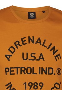 Camiseta de algodón naranja con cuello redondo y un audaz texto gráfico negro: "ADRENALINE U.S.A. PETROL IND. 1989" en un diseño circular.