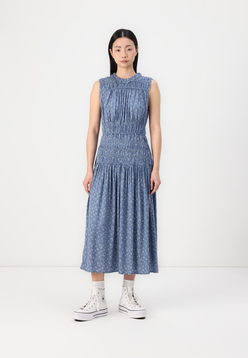 Blue floral cotton maxi dress with smocked bodice, textured ruffled neckline, and tiered skirt. Worn with white high-top sneakers.