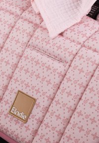 Rosa quiltat tyg med blomster- och fredssymbolmönster, med en tan-färgad Elodie-etikett och en vikta rosa musseltygdeka ovanpå.